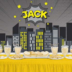May include: A yellow table is set with treats and decorations for a party. The backdrop features a cityscape with the name "JACK" in a comic book style. Glass jars and plates hold cookies and candies.