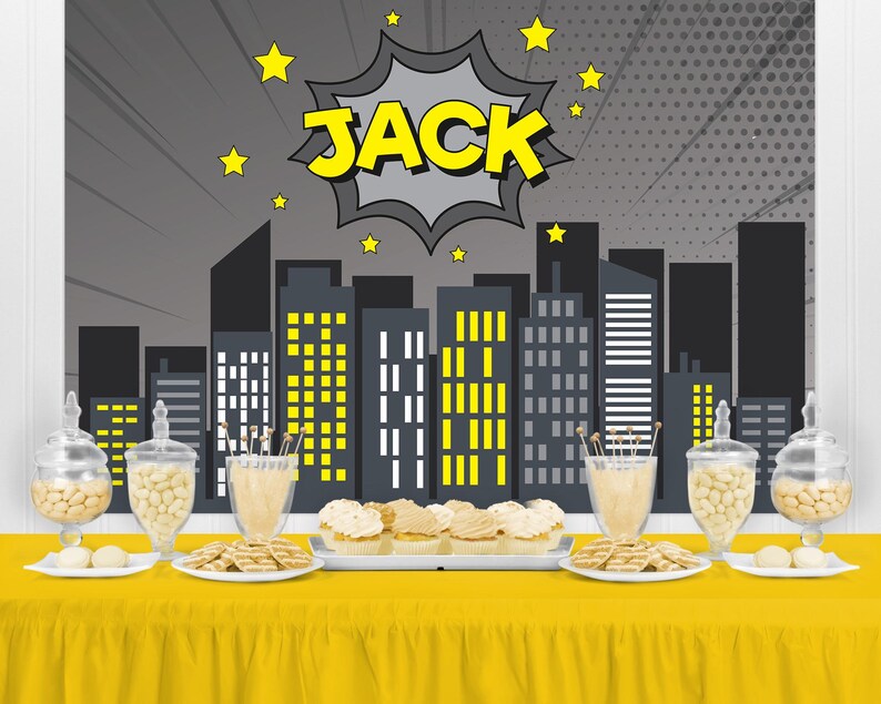 May include: A yellow table is set with treats and decorations for a party. The backdrop features a cityscape with the name "JACK" in a comic book style. Glass jars and plates hold cookies and candies.