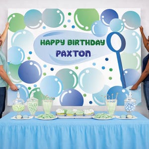 May include: A birthday party backdrop with a bubble theme. The backdrop features blue, green, and white bubbles with the text "HAPPY BIRTHDAY PAXTON". A blue table is set with treats and decorations. Balloons in pastel colors are on either side.