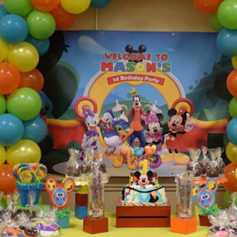 Puede incluir: Una escena de fiesta de cumplea&ntilde;os con una pancarta que dice "Welcome to Mason's 1st Birthday Party". Globos amarillos, verdes y naranjas rodean la escena. Una tarta con decoraci&oacute;n de Mickey Mouse adorna la mesa, junto con otros recuerdos de fiesta.