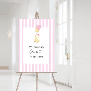 Puede incluir: Un letrero de bienvenida a rayas rosas y blancas sobre un caballete. El letrero presenta una ilustraci&oacute;n de acuarela de un patito amarillo sosteniendo globos. El texto dice "Bienvenida al 1er cumplea&ntilde;os de Charlotte."