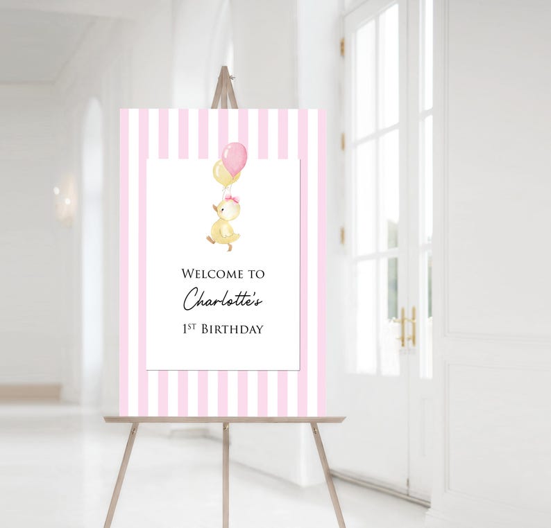 Puede incluir: Un letrero de bienvenida a rayas rosas y blancas sobre un caballete. El letrero presenta una ilustraci&oacute;n de acuarela de un patito amarillo sosteniendo globos. El texto dice "Bienvenida al 1er cumplea&ntilde;os de Charlotte."