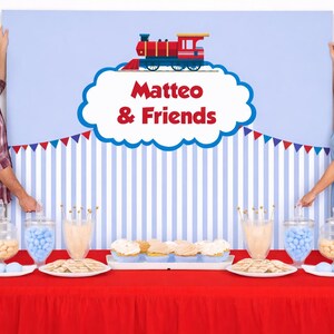 May include: A party scene with a red table, treats, and a backdrop featuring a train and the text "Matteo & Friends." Two people hold the backdrop. Balloons in pastel colors are on either side.