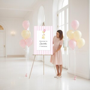 Puede incluir: Un letrero de bienvenida a rayas rosas y blancas sobre un caballete de madera, con el texto "Welcome to Charlotte's 1st Birthday". El letrero est&aacute; decorado con un pato de dibujos animados y globos. Globos rosas y amarillos atados al suelo.