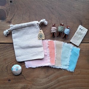May include: A set of craft supplies for making fabric flowers. The set includes a small bag, a button, three spools of thread, a piece of fabric with pins, and six fabric scraps in various shades of pink, peach, beige, blue, and turquoise.