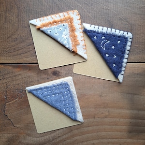 Pode incluir: Três marcadores de canto de tecido feitos à mão. Um marcador é azul com costuras brancas e um desenho de lua e estrelas. Outro marcador é azul com costuras brancas e um desenho floral. O terceiro marcador é laranja com costuras brancas e um desenho floral.