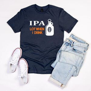 May include: Navy blue t-shirt with the text "IPA LOT WHEN I DRINK" and a white graphic of a beer bottle. The shirt is paired with light wash jeans and white trainers with red trim, all arranged on a white surface.