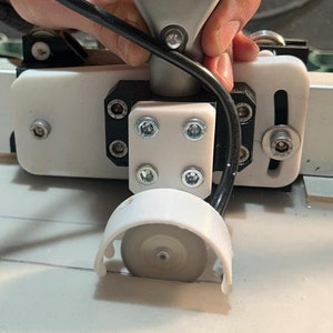 Może przedstawiać: Zbliżenie na żółto-szare narzędzie ręczne z przymocowanym czarnym kablem. Narzędzie jest zamontowane na białej plastikowej podstawie z małym białym kółkiem na dole. Narzędzie jest trzymane przez rękę.