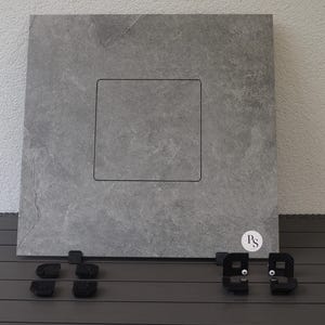 Op de afbeelding: Een grote, vierkante, grijze stenen tegel met een verzonken vierkant ontwerp in het midden. De tegel wordt geleverd met verschillende zwarte montagebeugels en een kleine, ronde sticker met de letters "PS".