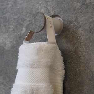 Peut inclure: Une serviette blanche et moelleuse est suspendue à un porte-serviettes chromé fixé à un mur de pierre grise. Le porte-serviettes a un design incurvé avec une sangle en tissu. La serviette est pliée et présente un motif texturé.