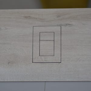 Può includere: Un pannello in legno grigio chiaro con un ritaglio rettangolare per un interruttore.
