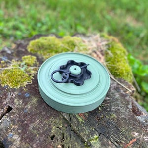 May include: A round, light green object with a black handle and a circular design. The object is sitting on a weathered tree stump with moss and grass in the background. The object appears to be a container or lid.