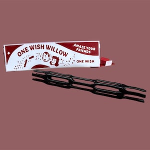 May include: A black, elongated, lattice-structured object positioned in front of a white rectangular box. The box features red text reading "ONE WISH WILLOW" and "AMAZE YOUR FRIENDS," along with a graphic illustration of two faces and a small star.