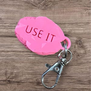 Puede incluir: Llavero rosa con un dije en forma de roca impreso en 3D que dice "USE IT" en letras rojas.