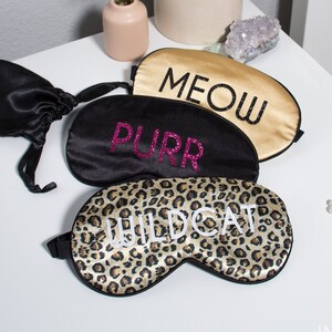 Op de afbeelding: Drie slaapmaskers met verschillende ontwerpen. De eerste is goudkleurig met het woord "MEOW" in zwart. De tweede is zwart met het woord "PURR" in roze glitter. De derde is een luipaardprint met het woord "WILDCAT" in wit.