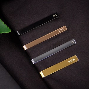 May include: Four tie bars in different colors and finishes: black, rose gold, silver, and gold. Each tie bar has a different engraved monogram: DS, RPT, TC, and ASM.