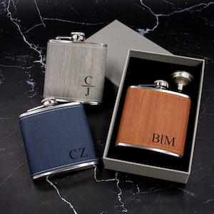 May include: Three personalized stainless steel hip flasks with different wood-grain finishes. The flasks are engraved with the initials "CJ", "BIM", and "CZ". One flask is in a gray box with a funnel.