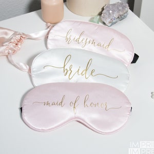 May include: Three satin sleep masks with gold lettering. The top mask is pink and says "bridesmaid". The middle mask is white and says "bride". The bottom mask is pink and says "maid of honor".