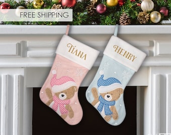 May include: Two personalized Christmas stockings with a teddy bear design. The stocking on the left is pink with a pink and white polka dot scarf and the stocking on the right is blue with a blue and white polka dot scarf. The stockings are hung on a white brick fireplace mantel with a green garland and red and gold ornaments.