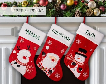May include: Three red Christmas stockings with white fur trim, hanging on a white brick fireplace mantel. The stockings are personalized with the names "Mama", "Christian", and "Papa". Each stocking features a different embroidered design: a snowman, Santa Claus, and a reindeer.