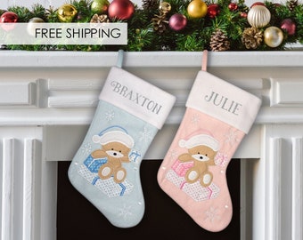 May include: Two personalized Christmas stockings with a teddy bear design. The stocking on the left is blue with the name "Braxton" embroidered on it. The stocking on the right is pink with the name "Julie" embroidered on it.