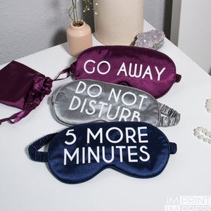 May include: Three satin sleep masks with different phrases printed on them. The top mask is purple and says "Go Away". The middle mask is silver and says "Do Not Disturb". The bottom mask is navy blue and says "5 More Minutes".