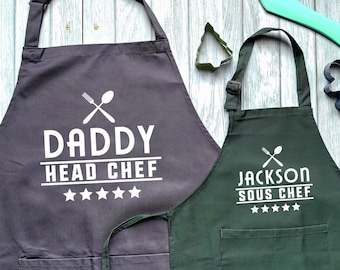 May include: Two black and green aprons with white text. The larger apron says "Daddy Head Chef" with five stars below the text. The smaller apron says "Jackson Sous Chef" with five stars below the text.