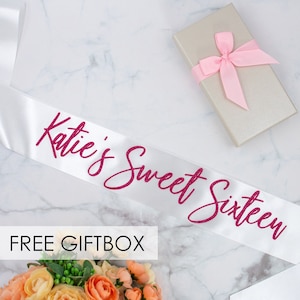May include: White satin sash with pink glitter lettering that reads "Katie's Sweet Sixteen". The sash is displayed on a white marble surface with a small gift box and flowers.