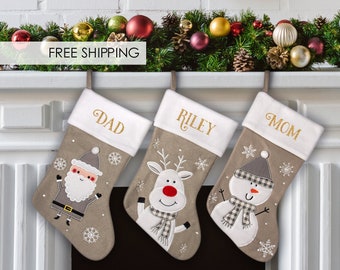 May include: Three personalized Christmas stockings with a Santa Claus, reindeer, and snowman design. The stockings are beige with white trim and gold lettering. The names "Dad", "Riley", and "Mom" are embroidered on the stockings.