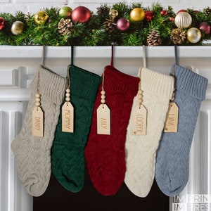 May include: Five knitted Christmas stockings with wooden tags hanging from a fireplace mantel. The stockings are beige, green, red, white, and gray. The tags are personalized with the names "Daddy", "Mommy", "Abby", "Cooper", and "Luna".