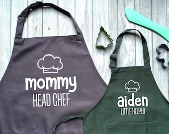 May include: Two kitchen aprons, one gray and one green, with white text. The gray apron says "mommy head chef" and the green apron says "aiden little helper". Both aprons have a chef hat graphic.