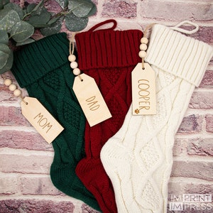 May include: Three knitted Christmas stockings with personalised wooden tags. The stockings are green, red, and white. The tags read "Mom", "Dad", and "Cooper".