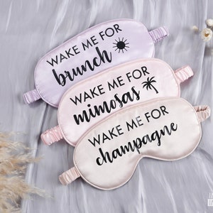 Puede incluir: Tres antifaces de satén con texto negro. El antifaz superior es lavanda y dice "Despiértame para el brunch". El antifaz del medio es rosa y dice "Despiértame para las mimosas". El antifaz inferior es rosa y dice "Despiértame para el champán".