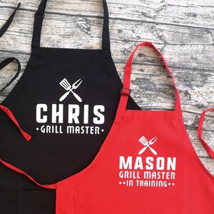 May include: Two black and red aprons with white text. The black apron says "Chris Grill Master" and the red apron says "Mason Grill Master - In Training".