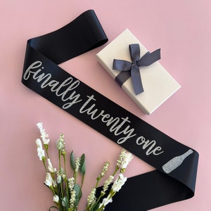 May include: Black satin sash with silver glitter lettering that reads "finally twenty one" and a silver glitter champagne bottle graphic.