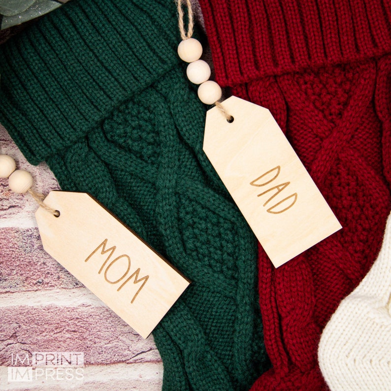 May include: Two knitted stockings with wooden tags that say "Mom" and "Dad". The stockings are green and red and have a cable knit pattern.