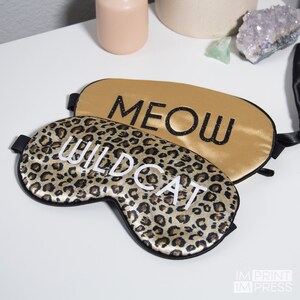 Op de afbeelding: Twee satijnen slaapmaskers met zwarte bies. Een masker is goudkleurig met het woord "MEOW" in zwart glitter. Het andere masker is bruin en zwart luipaardprint met het woord "WILDCAT" in wit.