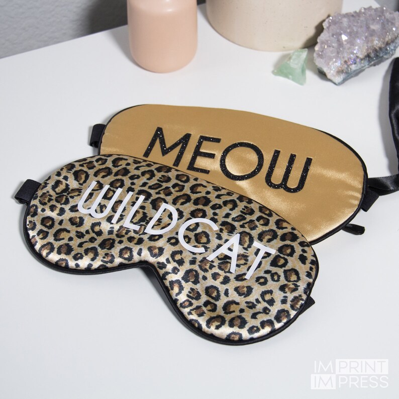 Op de afbeelding: Twee satijnen slaapmaskers met zwarte bies. Een masker is goudkleurig met het woord "MEOW" in zwart glitter. Het andere masker is bruin en zwart luipaardprint met het woord "WILDCAT" in wit.