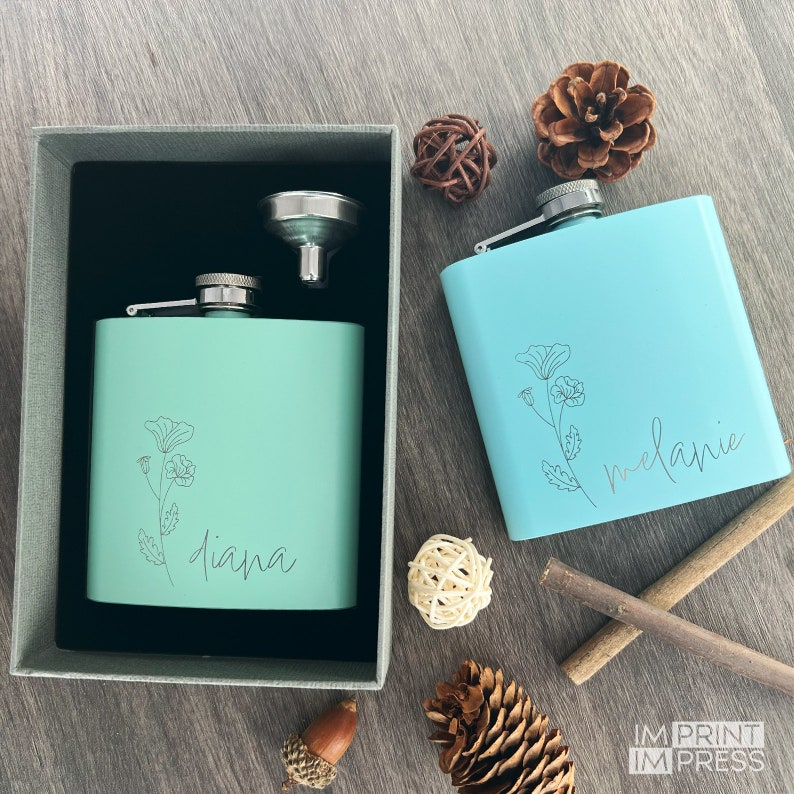 Personalized flask with flower and a name in a script font against a wood background surrounding by pine cones