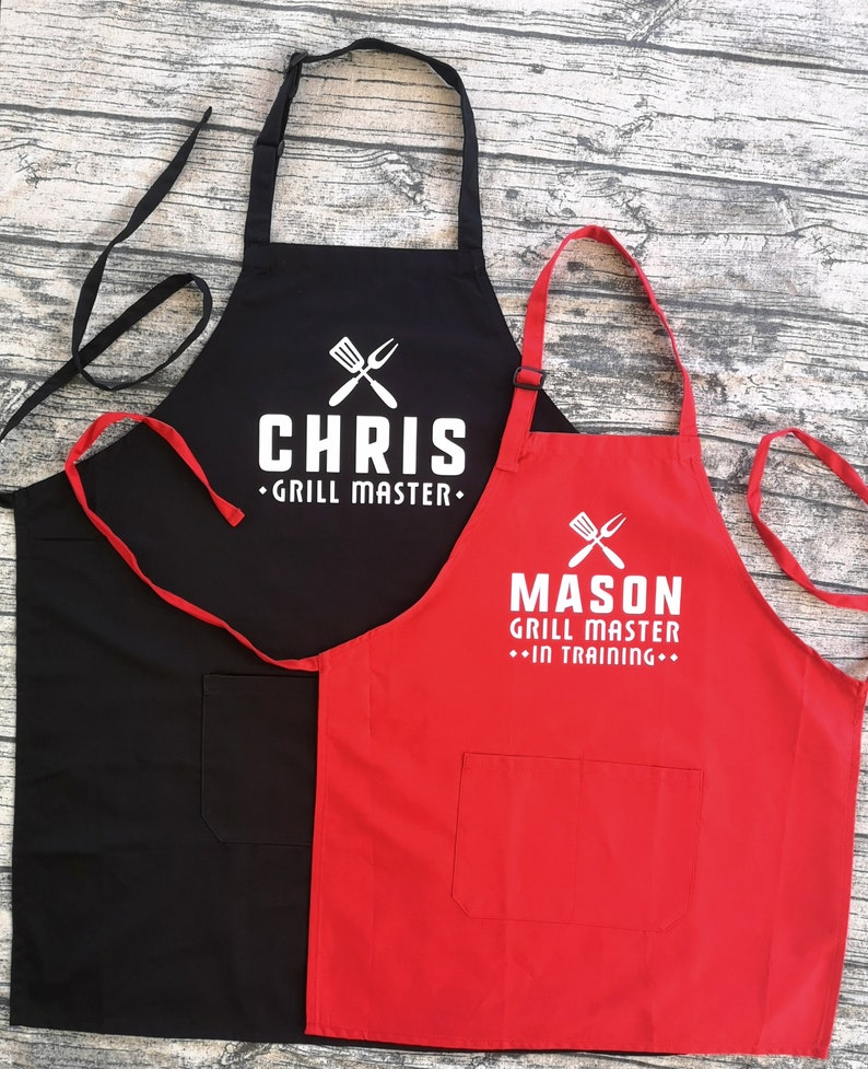 May include: Two black and red aprons with white text. The black apron says "Chris Grill Master" and the red apron says "Mason Grill Master - In Training".