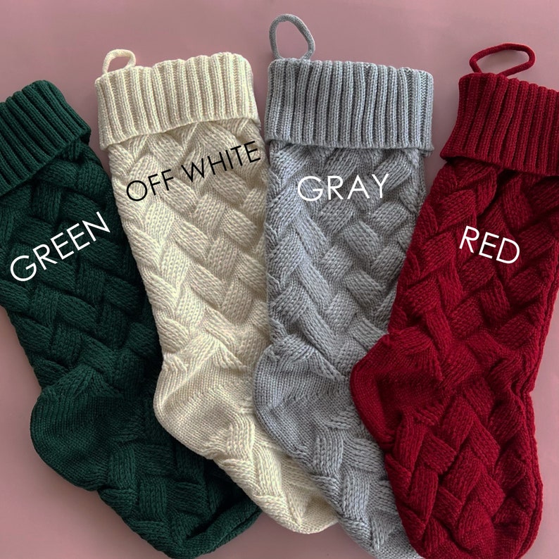 May include: Four knitted Christmas stockings in green, off-white, gray, and red. The stockings have a cable knit pattern and are hanging from a pink background.