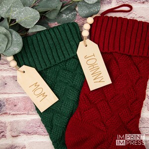 May include: Two knitted Christmas stockings with wooden gift tags. The green stocking has a tag that says "MOM" and the red stocking has a tag that says "JOHNNY".