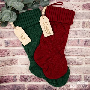 May include: Two knitted Christmas stockings with wooden tags. The red stocking has a tag that says "JOHNNY" and the green stocking has a tag that says "MOM".