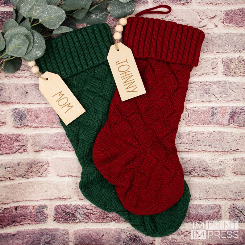 May include: Two knitted Christmas stockings with wooden tags. The red stocking has a tag that says "JOHNNY" and the green stocking has a tag that says "MOM".