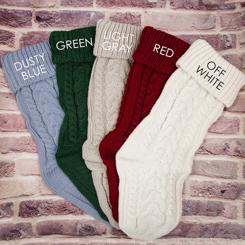 K&ouml;nnte beinhalten: F&uuml;nf gestrickte Weihnachtsstr&uuml;mpfe in verschiedenen Farben: staubiges Blau, Gr&uuml;n, Hellgrau, Rot und Off-White. Jeder Strumpf hat ein Zopfmuster und einen umgeschlagenen Bund mit dem Farbnamen darauf.