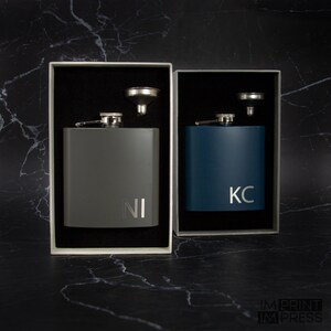 May include: Two personalized stainless steel hip flasks with funnels in gray and blue gift boxes. The flasks are engraved with the initials "NI" and "KC".