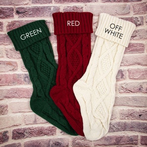 May include: Three knitted Christmas stockings in green, red, and off-white with a cable knit pattern. The stockings have the words "GREEN", "RED", and "OFF WHITE" written on the cuff.