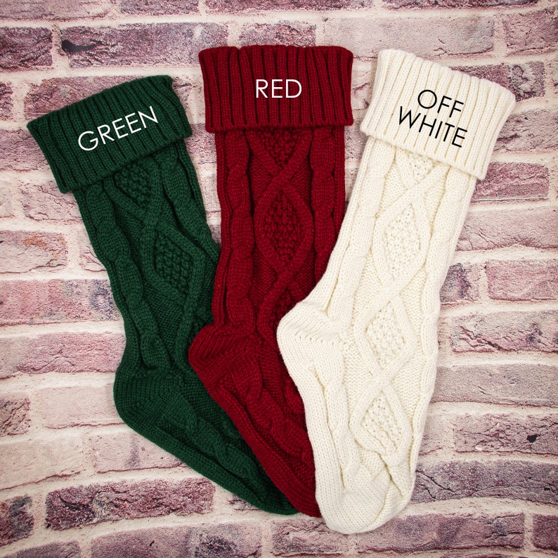 May include: Three knitted Christmas stockings in green, red, and off-white with a cable knit pattern. The stockings have the words "GREEN", "RED", and "OFF WHITE" written on the cuff.