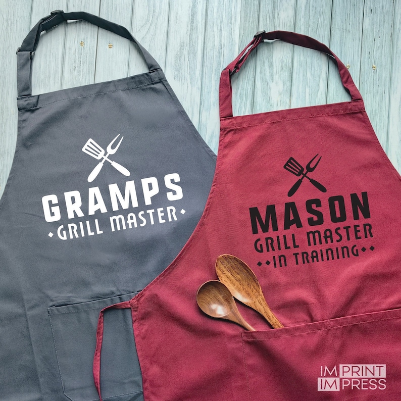 May include: Two gray and red aprons with a pocket and adjustable straps. The gray apron has white text that says "GRAMPS &bull; GRILL MASTER." The red apron has black text that says "MASON GRILL MASTER &bull; IN TRAINING &bull;".