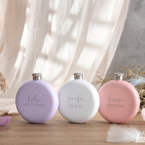 May include: Three round, personalized stainless steel hip flasks in lavender, white, and pink. The flasks are engraved with the names "Hallie Maid of Honor", "Jennifer The Bride", and "Karissa Bridesmaid".